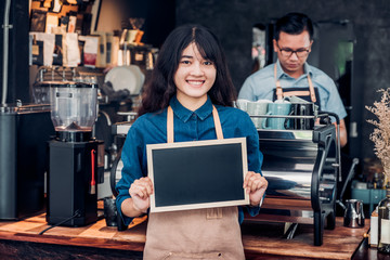 Poster - Asian female barista wear jean apron holding blank blackboard coffee menu at counter bar with colleague with smile face ,cafe service concept,business owner start upLeave space for adding your text