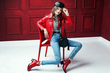 Full Body Studio Fashion Portrait Of Young Beautiful Woman Posing On Chair. Model Wearing Stylish Leather Cap, Biker Jacket, Stripped Turtleneck, Blue Jeans And Red Textured Ankle Boots. 