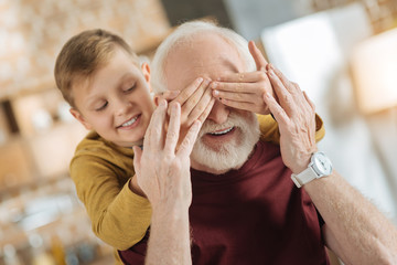 Wall Mural - Guess who. Positive cheerful nice boy standing behind his grandfather and closing his eyes while playing a guessing game with him