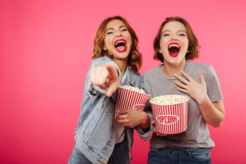 Wall Mural - Cheerful laughing women friends eating popcorn watch film pointing.