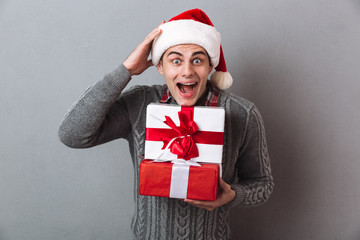 Shocked man in sweater and christmas hat holding gifts