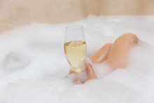 Close Up Photo Of Young Sexy Woman With Straight Naked Body And Clean Legs Lying And Relaxing In White Foam Bath Tub With Candles Around In Light Bathroom, Drink Alcohol From Wine Glass Indoors.