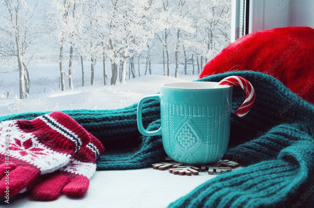 Winter background. Cup with candy cane, woolen scarf and red gloves on windowsill and winter forest outside - obrazy, fototapety, plakaty 