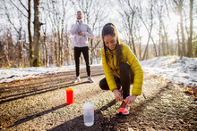 Close Up Of Sporty Active Beautiful Smiling Slim Woman In Sportswear Kneeling On The Road And Tying Shoelaces In The Sunny Winter Morning Outside In Nature With A Handsome Trainer Behind Her.