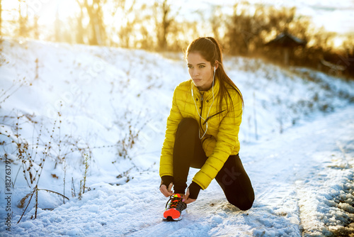 Fotografía Young sportive healthy focused girl tying shoelaces in winter sportswear on snowy winter road with earphones in the sunny morning