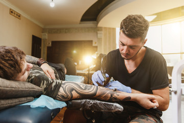 Professional tattooer artist doing picture on hand of man by machine black ink from a jar. Tattoo art on body. Equipment for making tattoo art. Master makes tattooed in light studio.