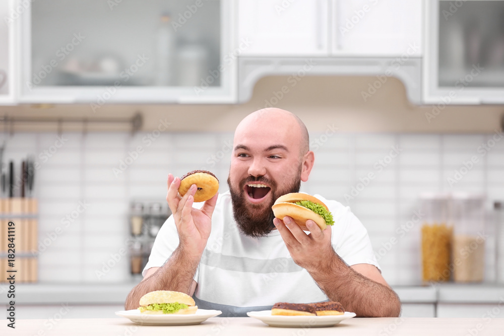 Overweight young man with unhealthy food at table in kitchen
