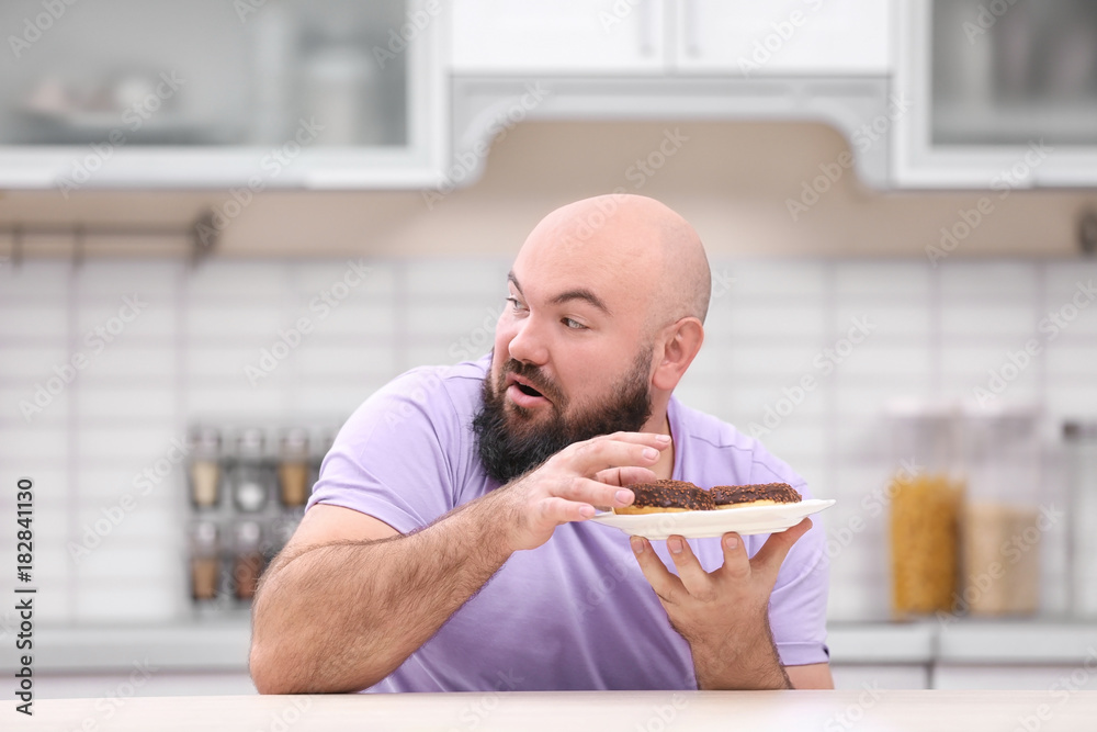 Overweight young man with unhealthy food at table in kitchen
