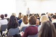© kasto - Business and entrepreneurship symposium. Female speaker giving a talk at business meeting. Audience in conference hall. Rear view of unrecognized participant in audience. Copy space on whitescreen.