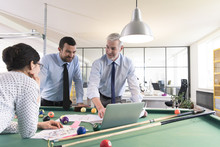Business People Standing At Pool Table With Laptop, Discussing Investment Strategy