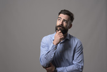 Young Adult Hipster Business Man Thinking And Looking Up At Copyspace While Touching Beard Against Gray Studio Background. 