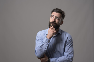 Young adult hipster business man thinking and looking up at copyspace while touching beard against gray studio background. 