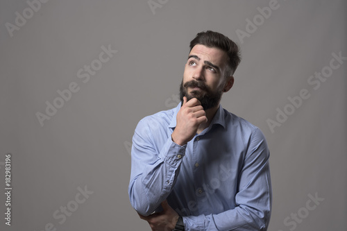 Cuadros en Lienzo Young adult hipster business man thinking and looking up at copyspace while touching beard against gray studio background