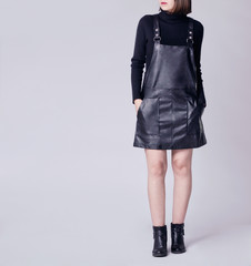 Poster - Young woman wearing stylish outfit with black faux leather pinafore dungaree mini dress, black turtleneck and black ankle boots isolated on light background. Copy space. Fashion details