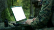 © Gorodenkoff - Close-up Soldier Uses Military Grade Laptop with White/Green Screen. In the Background Camouflaged Army Base in the Forest.