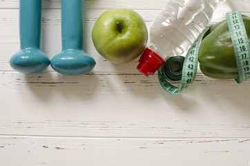 Wall Mural - Two dumbbells, bottle of water, green apple, centimeter on white wooden background with space  for text