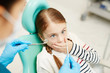 © pressmaster - Scared little girl covering her mouth by hand while looking at dental tools for oral check-up held by dentist