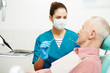 © pressmaster - Young dentist in protective mask, gloves and uniform holding hook and mirror and listening to senior patient
