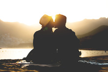Romantic Young Couple Kissing On The Beach On Sunset - Silhouette Of Teens Lovers At The Beginning Of Their Story Sitting On Sand - People, Love, Lifestyle, Relationship Concept