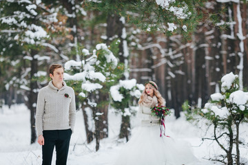 Wall Mural - Handsome groom in beige pullover on background of blurred bride in snowy forest. Winter wedding. Artwork.