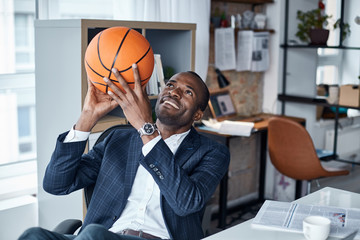 Playful mood. Cheerful young african manager in suit is sitting in office and holding orange basketball with smile. He is looking up while being ready to throwing it