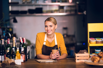 Wall Mural - Portrait of cheerful young woman worker tasting appetizing mug of tea while standing at counter in confectionary shop. Break concept