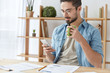 © wayhome.studio  - Busy young hardworking male sits at working desk, holds smart phone, view photos on Internet, drinks hot coffee or tea from green mug. People, technology and communication concept. Always in touch
