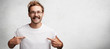 © wayhome.studio  - Horizontal shot of cheerful male with happy look points at white caual t shirt with copy space for your print or design. Bearded male nerd advertises new outfit, poses against blank white background