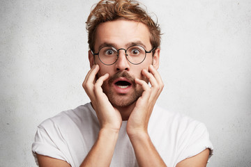 Wall Mural - Candid shot of stupefied shocked man stares at camera, wears round glasses, being in stupor as notices something awful and unexpected in front, doesn`t believe at what he sees. Surprisment concept