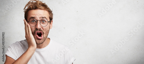 Cuadros en Lienzo Excited amazed bearded male keeps hand on cheek looks with joyful expression int
