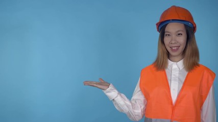Poster - young asian woman posing presenting products on blue background in studio . attractive millennial girl wearing white casual shirt and orange hard hat and vest looking at the camera with cheerful happy