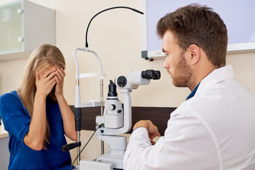 Portrait of young woman covering tired eyes sitting at slit lamp  during sight testing in optometrist office