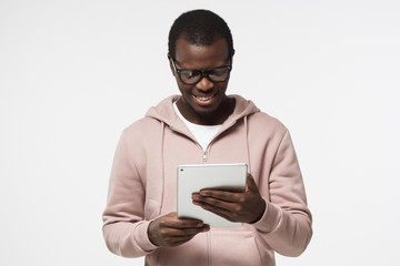 Wall Mural - Closeup portrait of young handsome African guy in hipster clothes isolated on white background looking attentively at screen of white tablet computer, experiencing digital opportunities of e-commerce