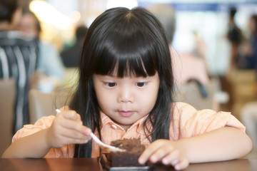 Wall Mural - Asian children cute or kid girl enjoy and fun with happy eating delicious brownie chocolate cake for sweet dessert or snack on wood table and make eyebalsl at lunch in restaurant or cafe