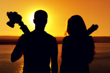 Rear View - Silhouettes Of Young Romantic Couple - Photographers Standing Together With Cameras And Tripod Against Background Of Sea Surface On Warm Sunny Day During Sunset
