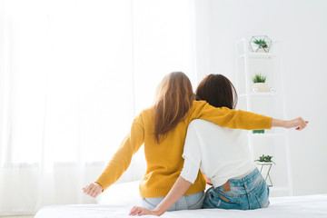 Wall Mural - Back view of women lesbian happy couple waking up in morning, sitting on bed, stretching in cozy bedroom, looking through window. Funny women after wakeup. Lesbian couple together indoors concept.