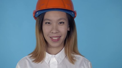 Aufkleber - close up portrait young asian architect wearing orange hard hat shows sign like on blue background in studio. attractive millennial girl looking at the camera