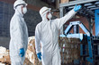 Portrait of two workers wearing biohazard suits standing in industrial warehouse of modern waste processing plant against recyclable cardboard in background