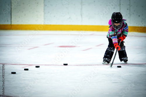 Fototapeta A kindergarten aged little girl in ski pants and winter jacket and helmet lining up a row of hockey pucks to practice shooting at a net