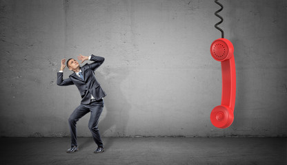 Wall Mural - a small scared businessman on concrete background near a large red retro phone handle hanging down.
