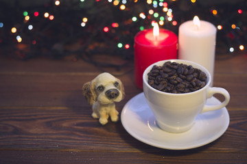 A romantic festive still life with a white cup filled with coffee beans, a toy puppy and two burning candles on a rustic wooden table. Dark blurred bokeh background