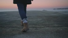 Close Up Slow Motion Shot Of Man Walk At Empty Sunrise Beach On Cold Winter Morning At Port Or Dock, Wears Sailor Themed Outfit, Winter Boots And Cosy Black Coat With Sweater
