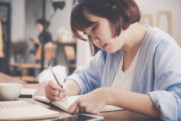 Young business woman in white dress sitting at table in cafe and writing in notebook. Asian woman talking smartphone and cup of coffee. Freelancer working in coffee shop. Vintage effect style pictures