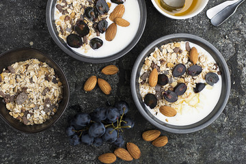 Wall Mural - Breakfast from healthy seasonal ingredients: granola, flakes, honey, dark grapes, almonds in a serving gray pial on a gray grunge background. Top view. Vegetarian Organic Food Concept.