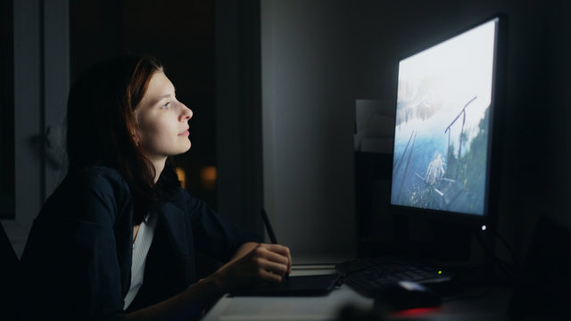 Concentrated young woman designer working at home at night using computer and graphics tablet to finish job