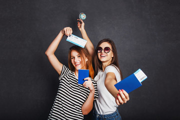 Cheerful happy girls holding passports, plane ticket and globe before grey background, indoor travel concept