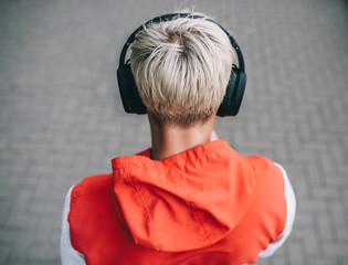 Rear view of blond young Caucasian guy walking down the street,  listening to music outside. Handsome man wearing red sportwear and black bluetooth headphones on head. People, technology concept.