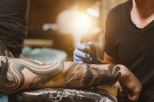 Close Up Of Professional Tattooer Artist Doing Picture On Hand Of Man By Machine Black Ink From A Jar. Tattoo Art On Body. Equipment For Making Tattoo Art. Master Makes Tattooed In Light Studio.