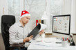 © ViDi Studio - Handsome serious and engrossed business man in shirt and red Christmas hat sitting at the desk, working at computer with modern monitor, with documents in light office on window background. New year.
