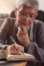 Senior Old Woman Writing Down Letters On A Piece Of Paper, Recording A Journal Or Diary Entry Or Writing A Novel